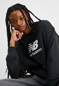 Zwarte sweatshirt met een wit New Balance-logo, met ribgebreide manchetten en een relaxte pasvorm. Model heeft lang vlecht haar en een neutrale achtergrond.