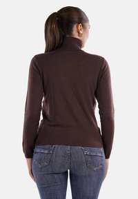 Maglione a collo alto marrone con maniche lunghe, design aderente e polsi ribattuti. Indossato con jeans in denim blu, mostrando un look casual e rilassato.