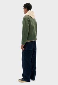 Man die zijwaarts kijkt, gekleed in een groene gewatteerde jas, een beige hoodie, brede donkerblauwe spijkerbroek met grote achterzakken en donkere sneakers.