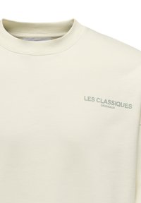 Helle beigefarbene Sweatshirt mit Rundhalsausschnitt, aus weichem Stoff gefertigt. Mit dezent in Grün aufgedruckt: "LES CLASSIQUES" und "ORIGINAUX".