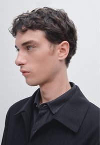 Czarny płaszcz z wyraźnie zarysowanym kołnierzem, noszony na ciemnej koszuli zapinanej na guziki. Falowane włosy, gładka skóra i neutralne tło podkreślają uwagę na ubraniu.