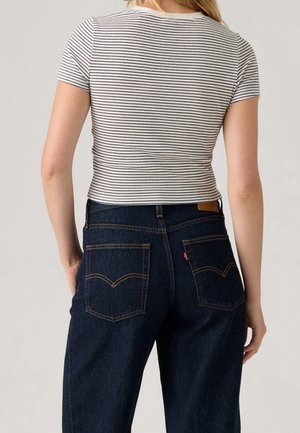 Vrouw met een wit-zwart gestreept shirt met korte mouwen, ingestopt in donkerblauwe hooggetailleerde spijkerbroek, gezien van achteren.