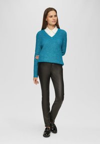 Türkiser Strickpullover mit V-Ausschnitt, kombiniert mit eng anliegenden schwarzen Lederhosen und schwarzen Schuhen. Der Pullover hat eine gerippte Textur und lange Ärmel.