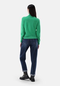 Grüner Strickpullover mit hohem Kragen, kombiniert mit locker sitzenden blauen Jeans und schwarzen Ankle-Boots, präsentiert eine lässige Rückansicht.