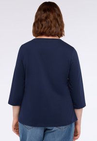 Frau mit schulterlangen braunen Haaren, die ein navyblaues Langarmshirt und eine blaue Jeans trägt, von hinten vor einem einfarbigen Hintergrund dargestellt.