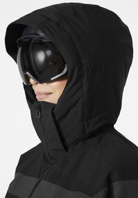 Černá izolační bunda s vysokým límcem a kapucí, se zipovým zapínáním a embosovaným logem, doplněná o elegantní lyžařské brýle.