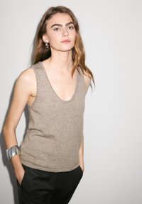 Lichtbeige mouwloze trui in zachte stof met een diepe V-hals. Model draagt een zwarte broek en een zilveren armband, wat een minimalistische stijl laat zien.
