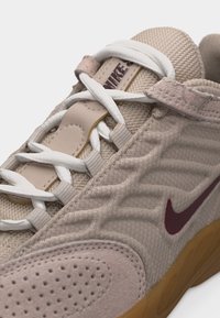 Μπεζ αθλητικό παπούτσι Nike με λευκά κορδόνια, υφή ύφασμα, διάτρητη σουέτ μύτη, μπορντό σήμα και ορατή καουτσούκ σόλα.