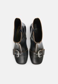 Botas de cuero negro hasta el tobillo con un acabado brillante, cremalleras laterales y un detalle de correa con hebilla. Presenta un pespunte marrón a lo largo de los bordes.