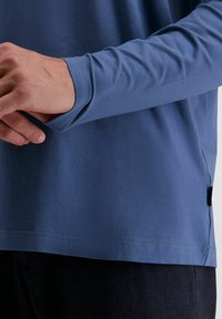 Mann, der den Ärmel seines blauen Langarmhemds anpasst, trägt dunkle Hosen und wird vom Taille bis zur Mitte des Arms vor einem neutralen Hintergrund gezeigt.