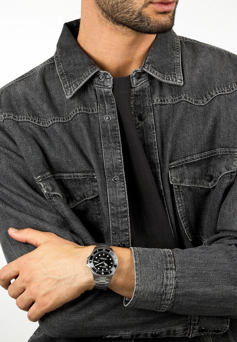 Giacca di jeans nera con ferramenta argentata, indossata sopra una camicia nera. Orologio d'argento con quadrante nero e indicatori luminosi al polso.