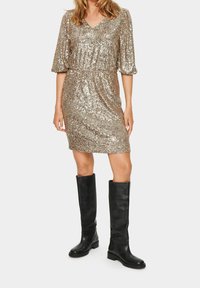 Guldklänning med paljetter och V-ringning, korta puffärmar och rynkad midja. Kombinerad med knähöga svarta läderstövlar.