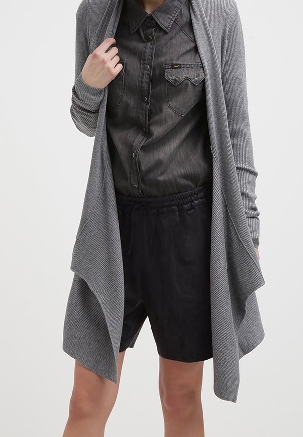 Femme portant une chemise boutonnée gris foncé, un short noir ample et un long cardigan gris ouvert sur le devant avec des manches côtelées.