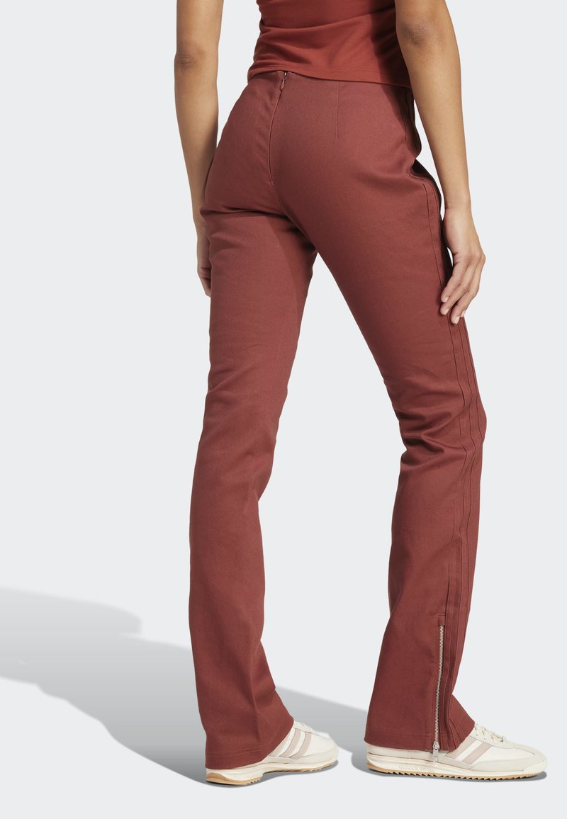 Personne portant un pantalon couleur rouille avec des fermetures éclair sur les côtés et des baskets blanches, debout de dos face à la caméra sur un fond uni.