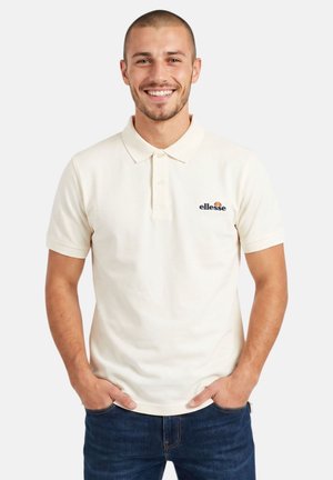Homme souriant aux cheveux courts, portant un polo blanc ellesse et un jean bleu, debout les mains dans les poches devant un fond blanc.