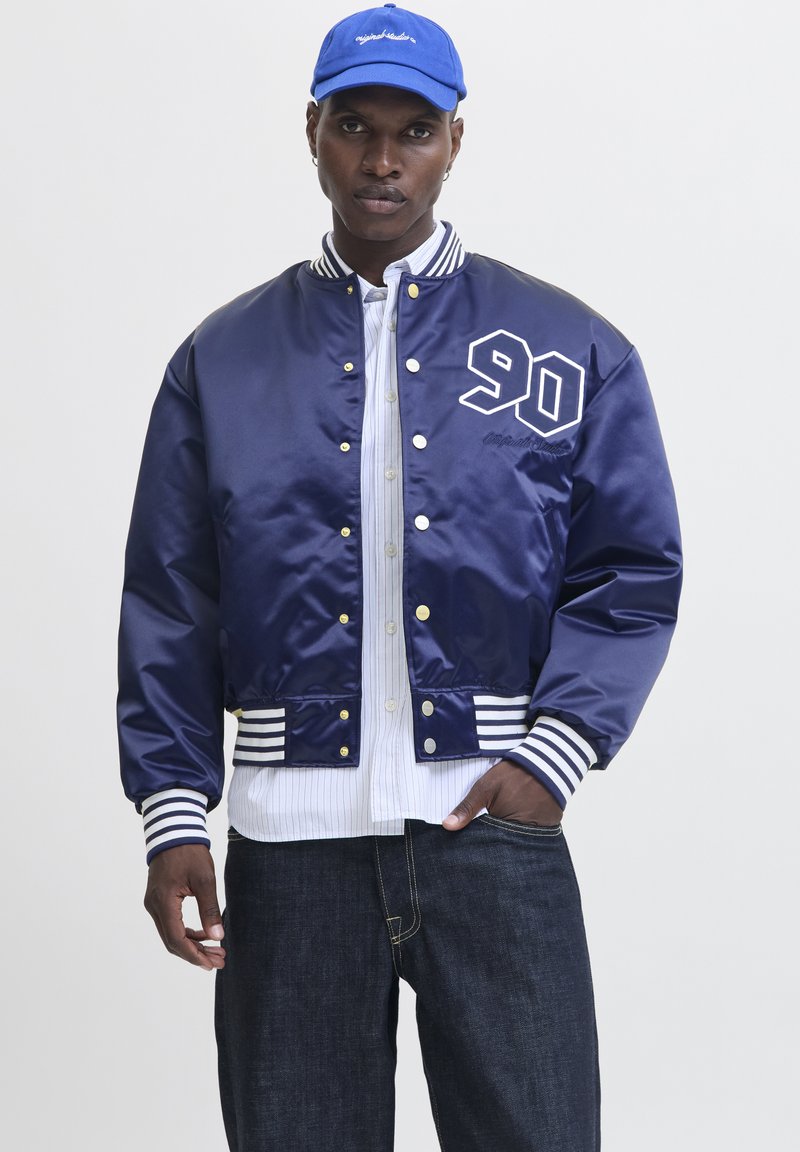 Homme portant une veste universitaire bleu marine avec poignets et taille rayés blancs, chemise blanche à boutons, jean foncé et casquette bleue.