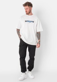 Witte katoenen T-shirt met grafische print; zwarte cargobroek met elastische boorden; witte sneakers. Model staat met de handen langs het lichaam tegen een effen achtergrond.