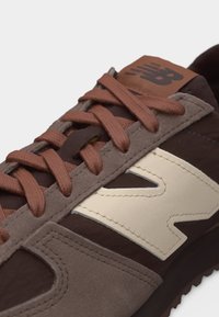 Gros plan d'une basket New Balance marron avec des détails en daim beige, des lacets marron et un grand logo "N" crème sur le côté.