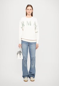 Biely sweatshirt s návrhom zeleného textu, v kombinácii s modrými džínsami so vzorom v tmavomodrej farbe a malou bielou kabelkou.