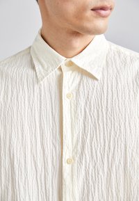 Homme portant une chemise boutonnée texturée de couleur blanc cassé avec un col pointu, visible du cou jusqu'à la partie supérieure de la poitrine, tourné de profil.