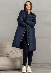 Marineblaue Pufferjacke mit langem, schlankem Design, gerippten Bündchen, Seitentaschen und silbernen Reißverschlüssen. Getragen über dunklen Hosen und Sneakers.