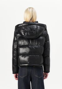 Schwarze Pufferjacke mit glänzender Oberfläche, ausgestattet mit einer Kapuze, horizontalem Steppdesign und elastischen Bündchen. Kombiniert mit dunklen Jeanshosen.