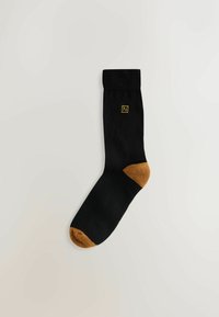Schwarze Socke mit goldenem Logo an der Seite, mit brauner Spitze und Ferse. Hergestellt aus glattem, gestricktem Material.