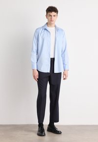 Light blue button-up shirt with long sleeves, worn over a white t-shirt, paired with dark trousers and black shoes. Simple design, no patterns.
