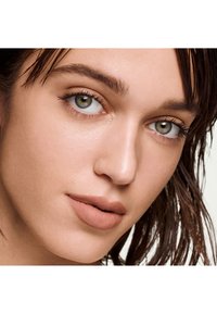 Gros plan d'un visage présentant une peau lisse et claire, des yeux verts avec des cils subtils et des lèvres nude mates, encadré par des cheveux bruns mouillés.