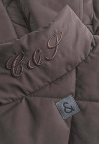 Braun gestepptes Material mit glatter Textur. Verfügt über braun gestickte Buchstaben "C G" und ein graues Stoffetikett mit einem "&"-Symbol.