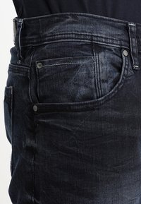 Närbild av en ficka på svarta denimjeans med synliga bältesslingor, nitar och sömnadsdetaljer på en person som bär en mörk skjorta.