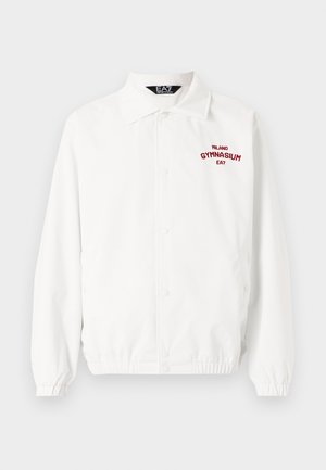 White coach jacket with a front button closure, elastic cuffs, and "MILANO GYMNASIUM EA7" embroidered in red on the chest.