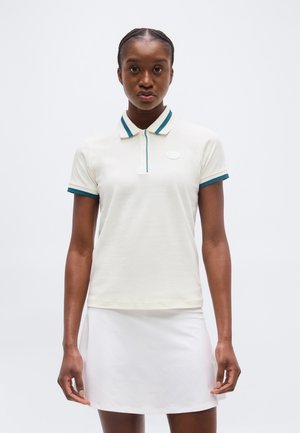 Žena oblečená v bielom polokošeli s krátkym rukávom a tyrkysovým lemom a bielej sukni, stojaca pred jednotným svetlým pozadím.