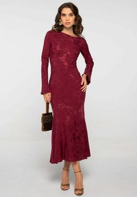 Vestido de encaje floral burdeos con mangas largas, dobladillo acampanado, textura suave y escote barco; accessorios con un pequeño bolso con flecos.