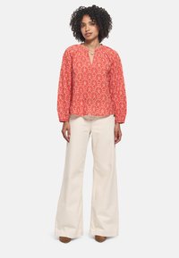 Blusa floral roja con mangas largas y abullonadas, y un escote de media abertura, combinada con pantalones de pierna ancha en color crema y zapatos de tacón marrones.