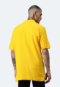 Gelbes Poloshirt aus Baumwolle, kurze Ärmel und seitliche Schlitze. Auffällige Tattoo-Details an den Armen, von hinten gezeigt.