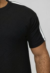 T-shirt noir texturé avec des manches courtes, un col rond et des rayures blanches le long des coutures des épaules.