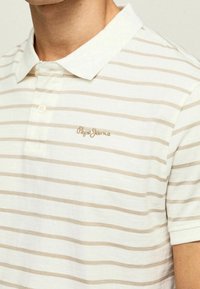 Stribet polo i lysecreme med beige vandrette striber. Har en knappet krave og broderet logo på brystet. Bomuldsmateriale.