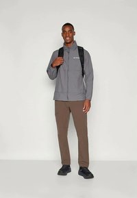 Graue leichte Jacke mit Kapuze, schwarzer Rucksack, braune Hose und schwarze Sneaker. Einfaches Design und lockere Passform, die sich für Outdoor-Aktivitäten eignet.