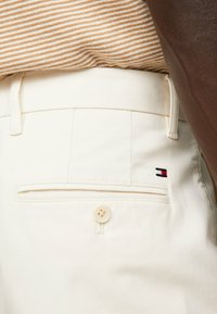 Pantaloni in cotone bianchi con una tasca posteriore, caratterizzati da un piccolo logo rosso, bianco e blu. Include un bottone beige e dettagli cuciti.