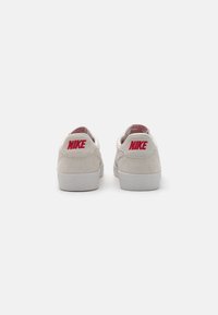 Nike tenisice od svijetlosmeđeg pliša s crvenim akcentima na logu. Niska verzija, s mekanim ovratnikom i hrapavim bijelim gumama.