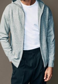 Lichtgrijze rits-hoodie met een zachte textuur, gedragen over een wit T-shirt. Gecombineerd met donker gestreepte broeken, ontworpen voor comfort.