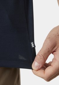 Námořnická modrá košile s krátkým rukávem vyrobená z měkké látky, s malou bílou nášivkou na lemu. Lem má mírný rozparek.