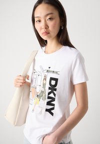 Baltos medvilnės marškiniai su grafiniu moters atvaizdu ir tekstūra išryškintais "DKNY" rašmenimis. Modelis laiko šviesios spalvos rankinę.