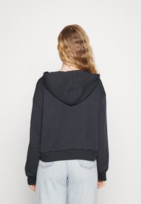 Hoodie corto nero realizzato in tessuto morbido, con cappuccio regolabile e polsini a costine. Il retro presenta una costruzione senza cuciture e una vestibilità casual.