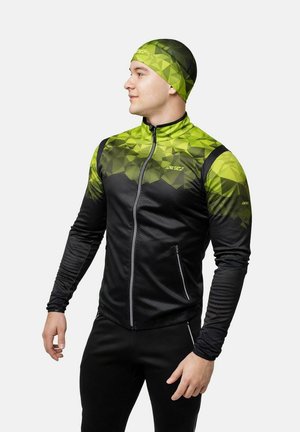 Uomo che indossa una giacca sportiva con motivi geometrici neri e verdi e un cappellino abbinato, in piedi con postura rilassata, che guarda di lato.