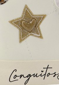 Zapatilla color crema con un acento de estrella dorada texturizada con un corazón, delineado con costuras, y el nombre de la marca "Conguitos" en letra cursiva negra en la base.