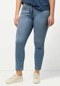 Jeans in denim blu con un design aderente, dotati di una cintura elastica e decorazioni sottili vicino alla tasca anteriore; indossati con scarpe da ginnastica bianche.