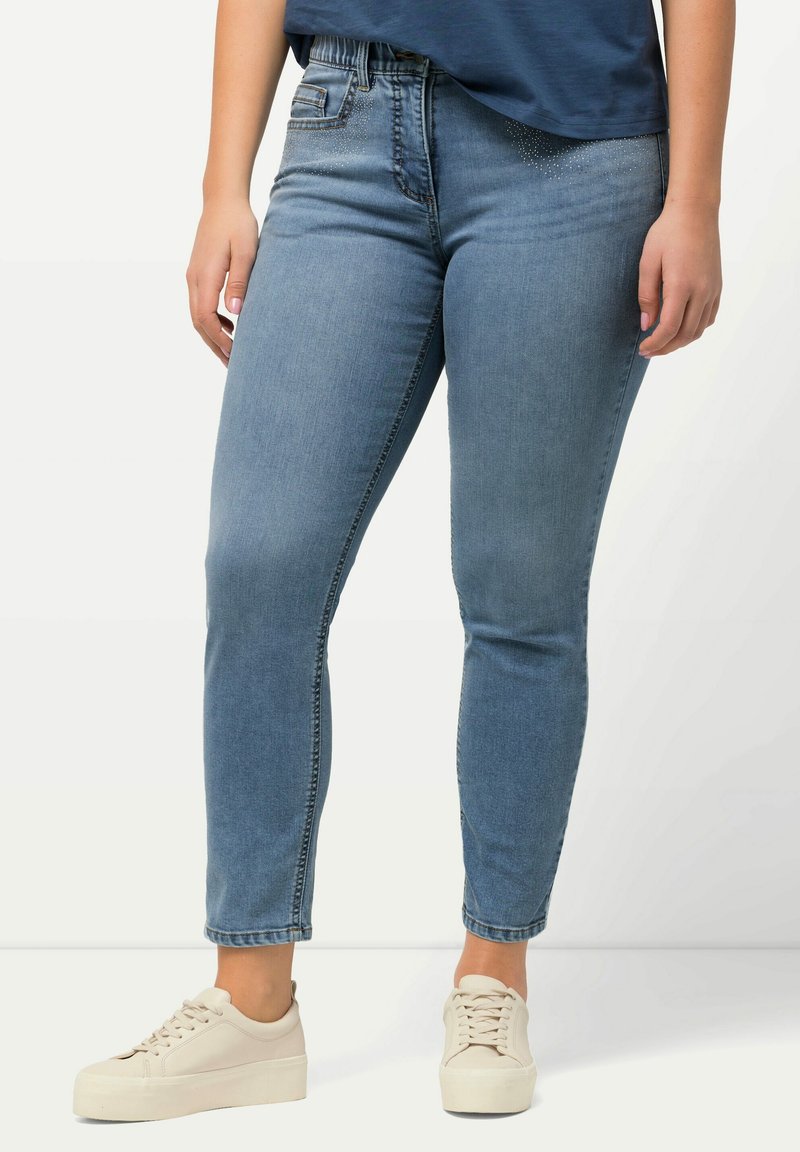Jeans in denim blu con un design aderente, dotati di una cintura elastica e decorazioni sottili vicino alla tasca anteriore; indossati con scarpe da ginnastica bianche.