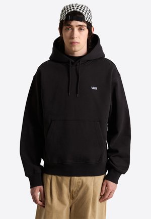 Giovane uomo con felpa con cappuccio Vans nera, pantaloni beige e cappellino a quadri nero e bianco indossato al contrario, in piedi davanti a uno sfondo semplice.